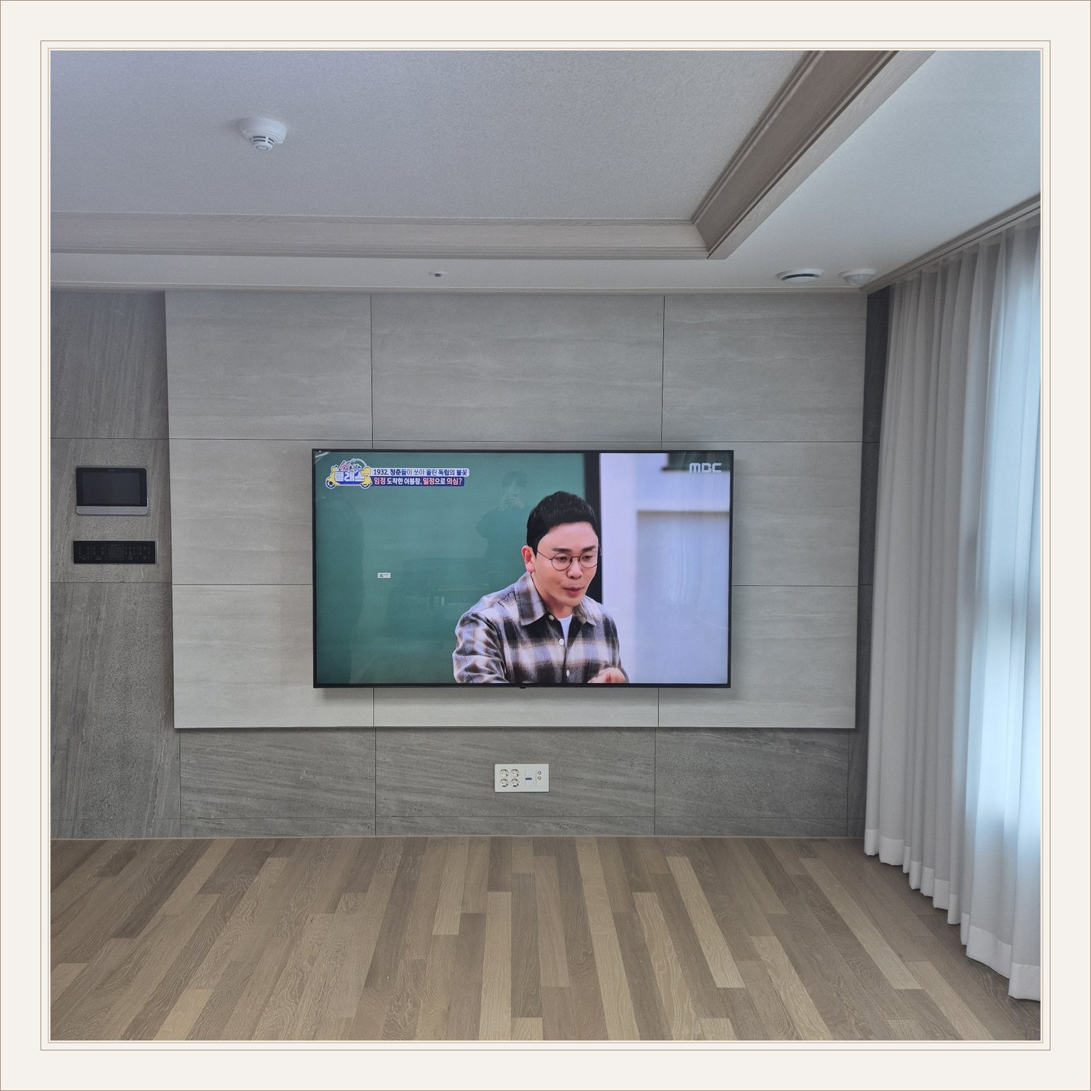 거실 회색 패널과 벽걸이 TV, 오른쪽 커튼 옆으로 화면이 중심에 놓여 있습니다