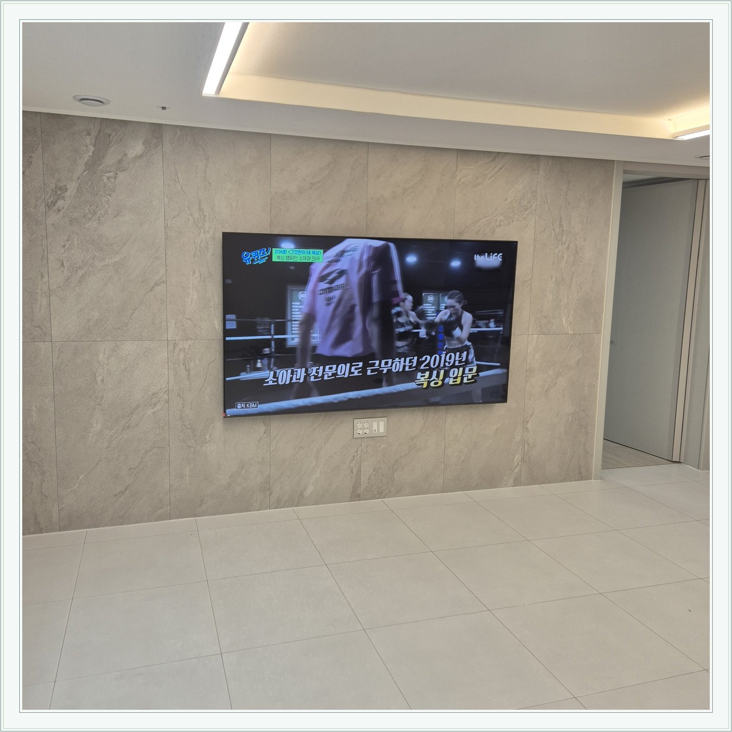 석재 느낌 벽면을 정면에서 본 TV, 넓은 바닥 여유와 조명 라인이 동시에 보입니다