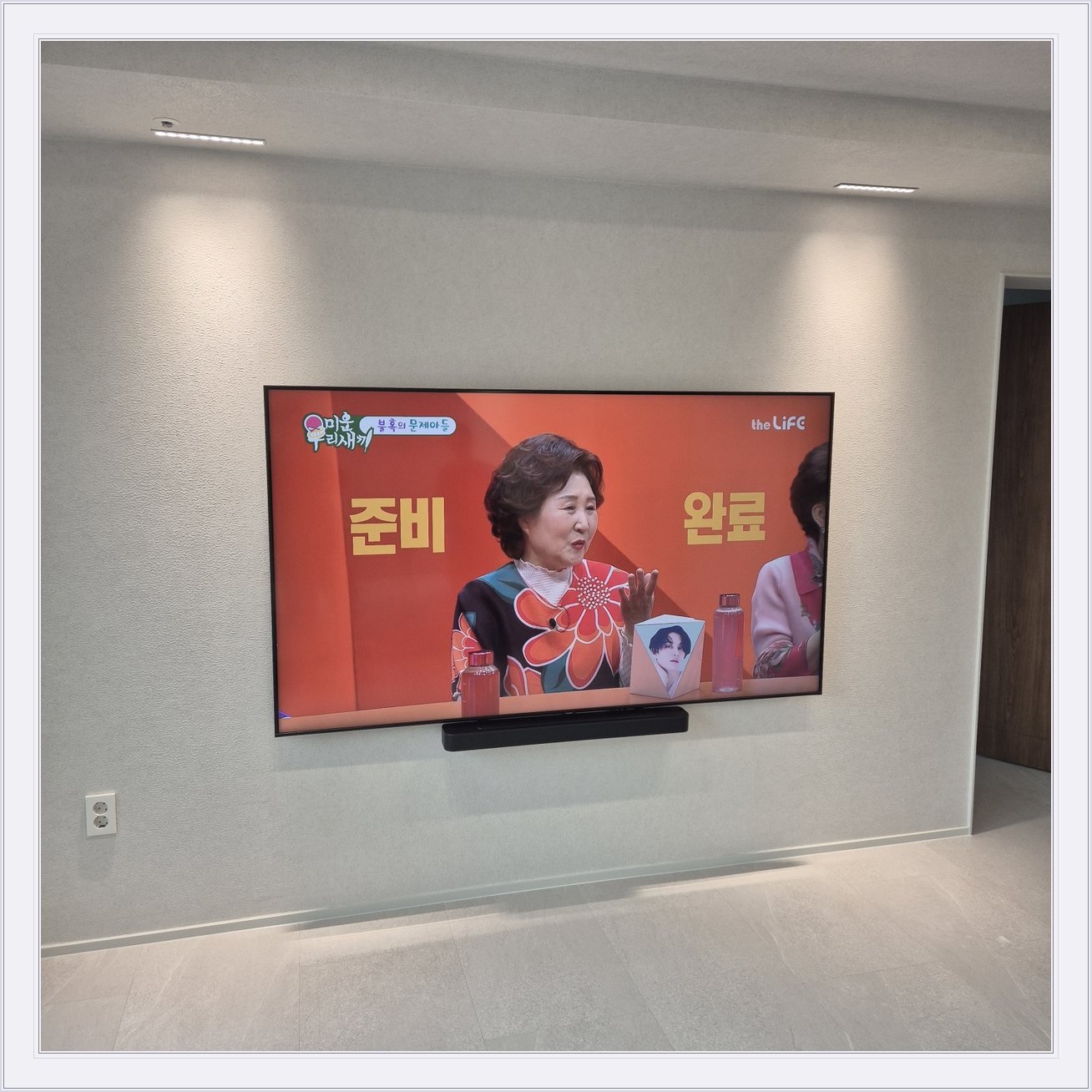 조명이 비추는 미색 벽과 사운드바, 화면 아래 가로로 놓인 검은 음향기기가 선명합니다