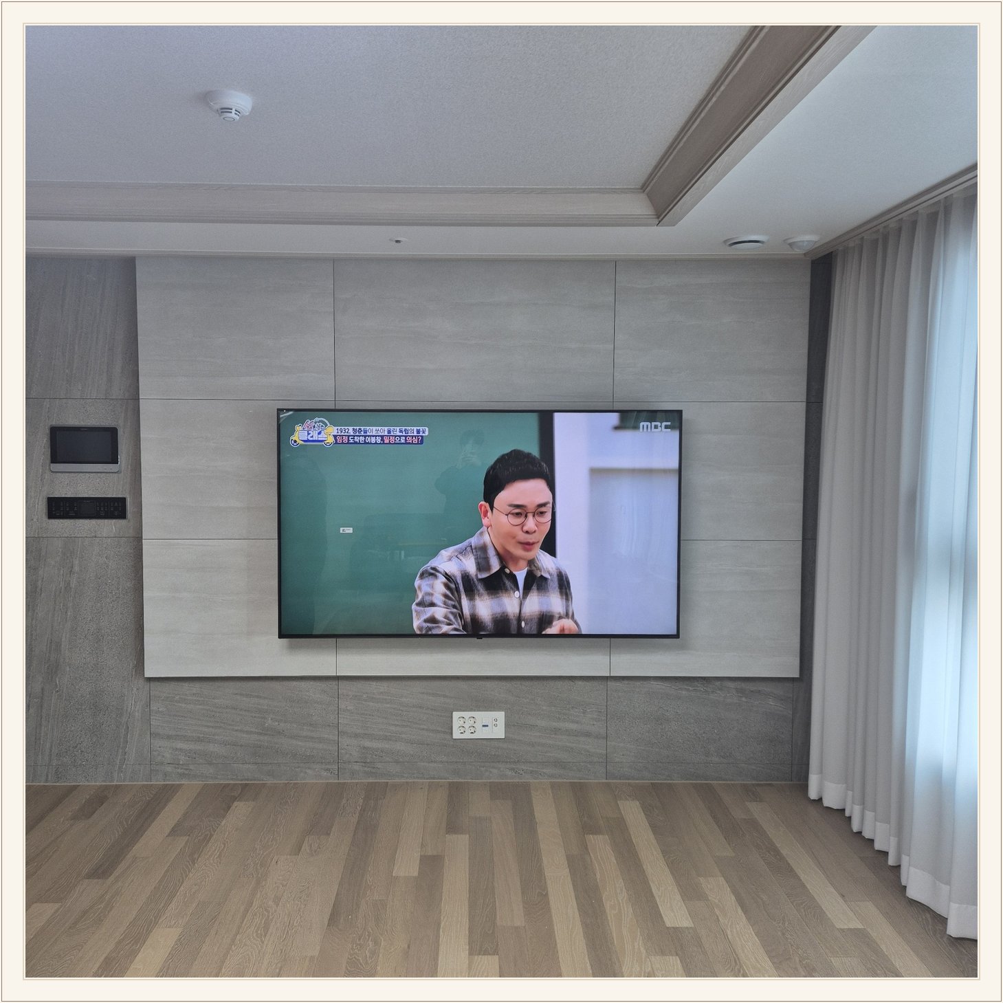 거실 회색 패널과 벽걸이 TV, 오른쪽 커튼 옆으로 화면이 중심에 놓여 있습니다
