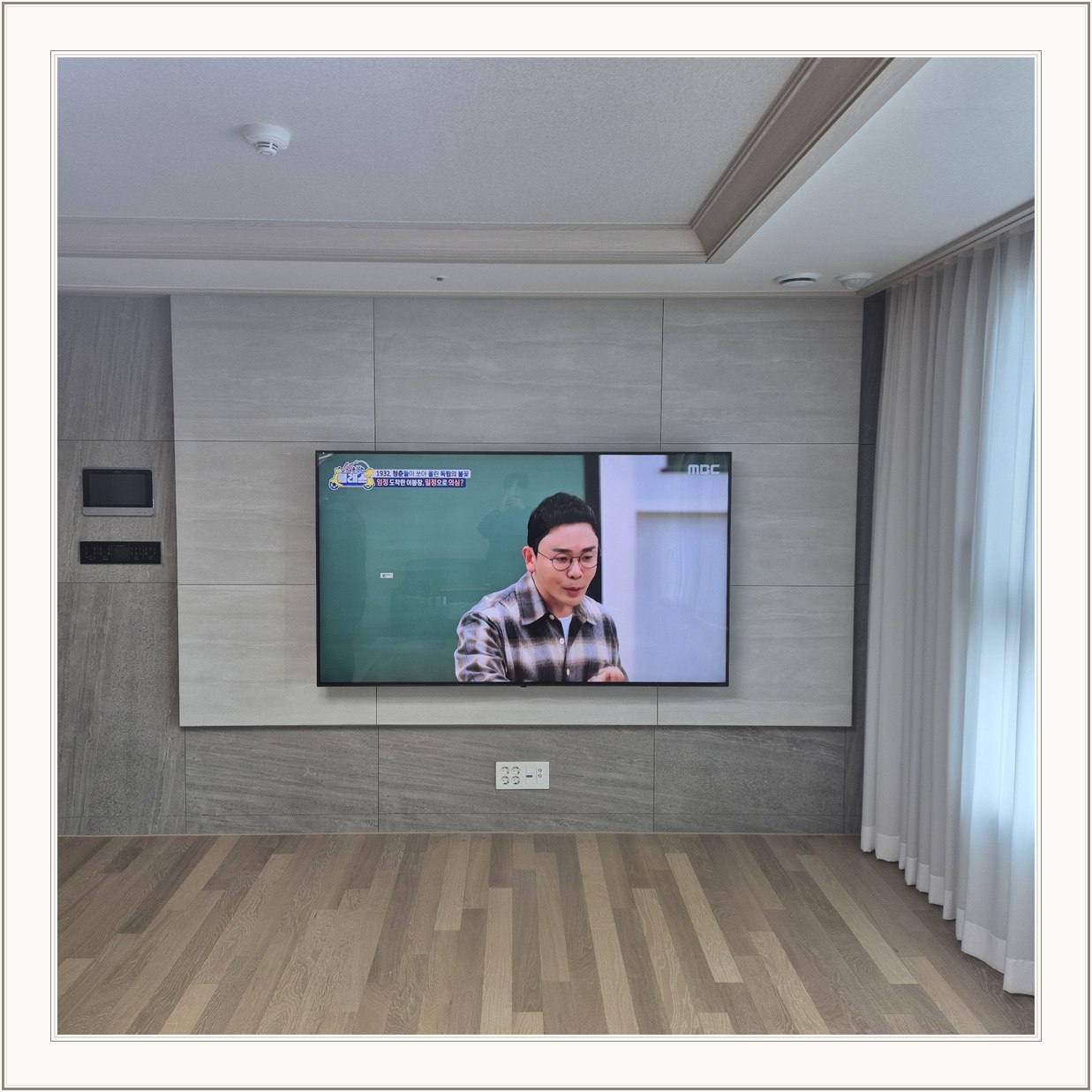거실 회색 패널과 벽걸이 TV, 오른쪽 커튼 옆으로 화면이 중심에 놓여 있습니다