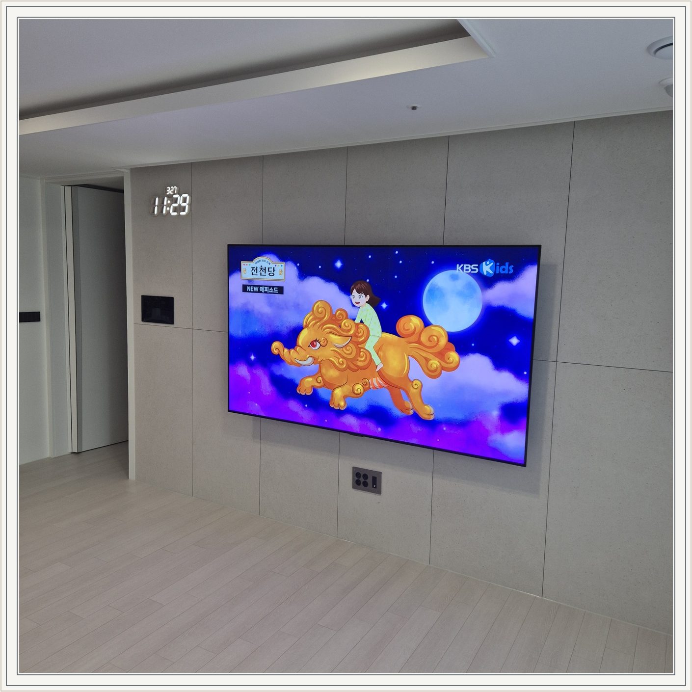 회색 판넬 벽 위의 화면과 디지털 시계, 벽면 패널 이음선과 TV 중심의 배치를 비교할 수 있습니다