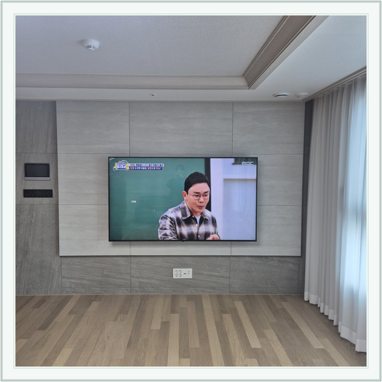 거실 회색 패널과 벽걸이 TV, TV 아래 콘센트와 양쪽 마감 판넬의 대비가 뚜렷합니다