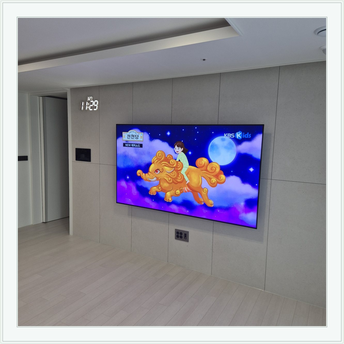 회색 판넬 벽 위의 화면과 디지털 시계, 벽면 패널 이음선과 TV 중심의 배치를 비교할 수 있습니다